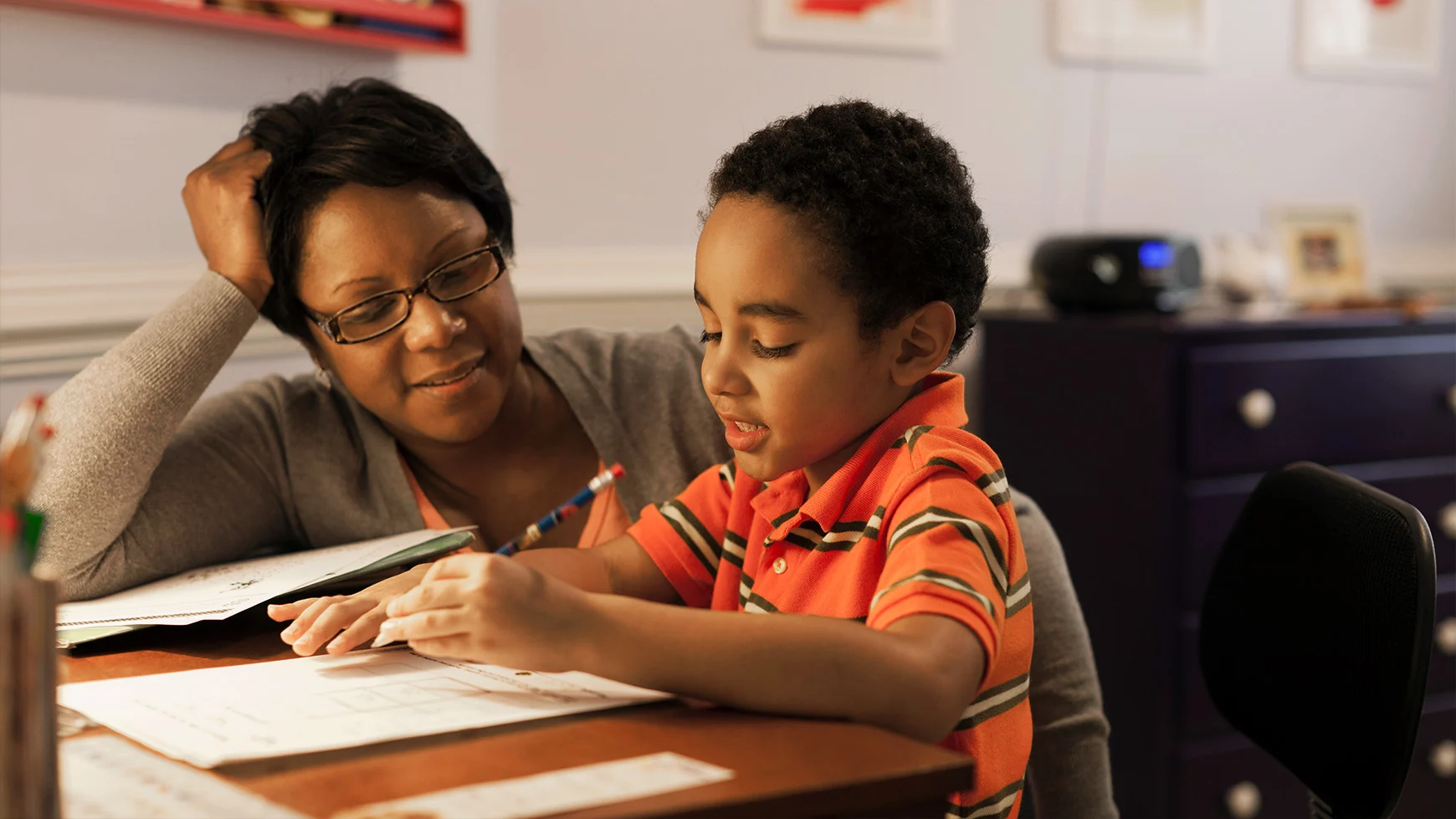 A parent sits next to their child, watching as the child, holding a pencil, works on a writing assignment. They both smile.