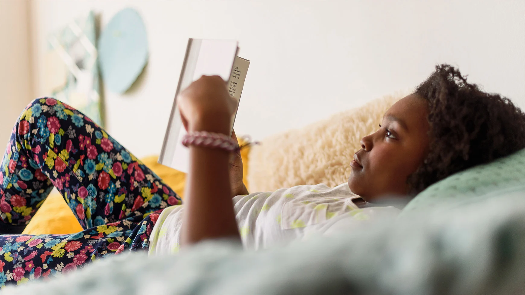 A child reading a book while lying down, wearing flowered pants and a white t-shirt.